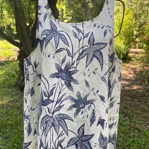 May include: A sleeveless tank top in off-white fabric featuring a dark blue botanical print. The pattern displays various large star-shaped flowers, slender leaves, and small butterfly illustrations distributed across the entire garment surface.