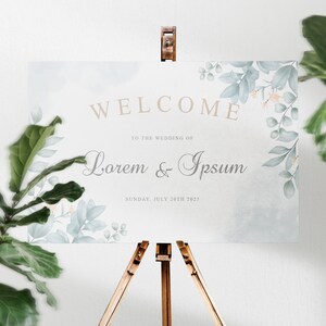 K&ouml;nnte beinhalten: Ein Aquarell-Willkommensschild f&uuml;r eine Hochzeit mit einem floralen Design in Blaut&ouml;nen und Gr&uuml;n. Das Schild lautet "Willkommen zur Hochzeit von Lorem & Ipsum, Sonntag, 30. Juli 2023".