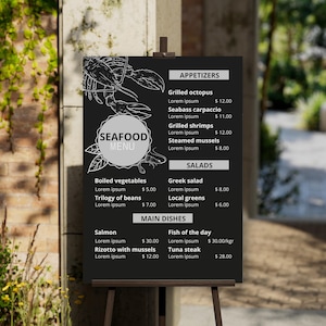 K&ouml;nnte beinhalten: Eine schwarze Tafel mit wei&szlig;er Schrift, die Meeresfr&uuml;chtegerichte und Preise auflistet. Die Speisekarte tr&auml;gt den Titel "Seafood Menu" und umfasst Vorspeisen, Salate und Hauptgerichte. Einige der aufgef&uuml;hrten Gerichte sind gegrillter Tintenfisch, Seehecht-Carpaccio, gegrillte Garnelen, ged&auml;mpfte Muscheln, gekochtes Gem&uuml;se, Bohnentrio, griechischer Salat, gr&uuml;nes Gem&uuml;se, Lachs, Fisch des Tages, Risotto mit Muscheln und Thunfischsteak.