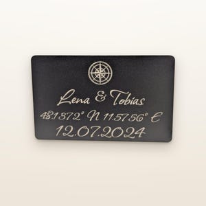 Könnte beinhalten: Eine rechteckige schwarze Platte mit einem eingravierten Kompassrosen-Symbol oben. Der Text lautet Lena & Tobias, gefolgt von den Koordinaten 48.1372° N 11.5756° E und dem Datum 12.07.2024, alles in einer hellen Schreib- und Sans-Serif-Schrift.
