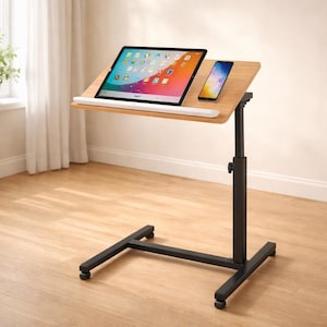 May include: A mobile laptop desk with a light wood-grain surface, holding a tablet and smartphone. The desk has a black, adjustable-height frame with a wheeled base, designed for use in a home or office setting.