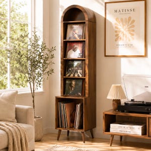 May include: A tall, wooden record storage cabinet with an arched top. The cabinet has multiple shelves displaying vinyl records. The bottom section holds additional records. The cabinet is in a well-lit room with a window, a sofa, and a side table.