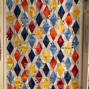 Könnte beinhalten: Ein rechteckiger Quilt mit einem sich wiederholenden Rautenmuster aus verschiedenen bunten Stoffresten in Rot-, Orange-, Gelb-, Blau- sowie Schwarz-Weiß-Drucken. Der Text patcherymenagerie.blogspot.com befindet sich am unteren Rand.