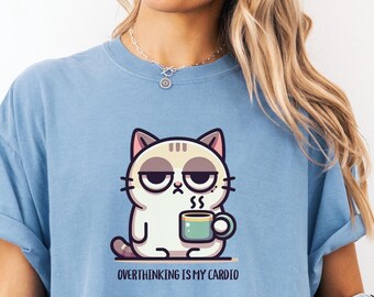 May include: Light blue t-shirt featuring a cartoon cat holding a mug. The cat has a grumpy expression and the text "Overthinking is my cardio" is printed below the cat. The shirt has short sleeves and a relaxed fit.