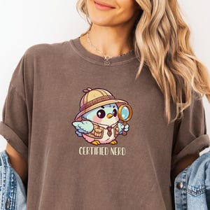 以下が含まれることがあります： 茶色のTシャツ。漫画の鳥がサファリハットをかぶり、虫眼鏡を持っています。鳥の下には「CERTIFIED NERD」の文字がプリントされています。Tシャツはデニムジャケットと合わせて着用されています。