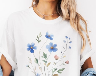 May include: White t-shirt featuring a watercolor floral design with blue and pink flowers, green leaves, and stems. The shirt is paired with a denim jacket and shorts. The overall aesthetic is casual and feminine.