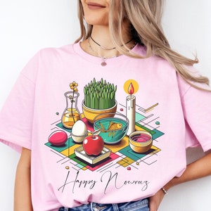 May include: Pink t-shirt with a graphic design of a traditional Persian New Year (Nowruz) Haft Seen table setting. The design features a bowl of wheatgrass, a goldfish bowl, a red apple, a candle, a coin, and a decorated egg. The text "Happy Nowruz" is printed below the design.