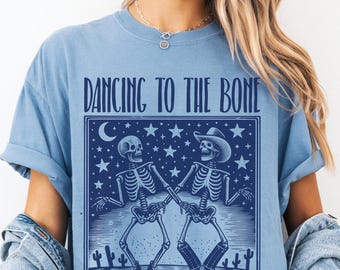 May include: A pale blue t-shirt featuring the words "DANCING TO THE BONE" above a dark blue graphic of two dancing skeletons. The graphic includes stars, a crescent moon, and cacti. The shirt is worn by a person.