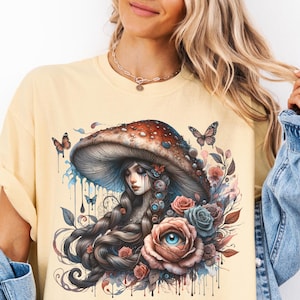 May include: A yellow t-shirt with a graphic of a woman with long dark hair under a mushroom with red and brown accents. The mushroom is surrounded by blue and pink flowers. The graphic has a watercolor effect with dripping paint.