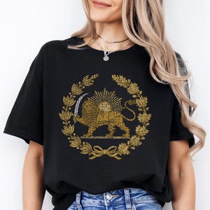 Könnte beinhalten: Schwarzes T-Shirt mit einem goldenen Löwen- und Sonnenemblem in einem Lorbeerkranz. Das Design ist auf dem T-Shirt zentriert, mit einer detaillierten Illustration des Löwen, der ein Schwert hält. Das T-Shirt wird mit blauen Jeans kombiniert.