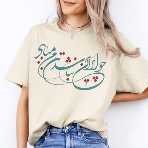 Op de afbeelding: Crèmekleurig T-shirt met turquoise en kastanjebruine kalligrafie. Het shirt wordt gecombineerd met een blauwe spijkerbroek. De tekst op het shirt is in een elegant schrift. De algehele stijl is casual en comfortabel.