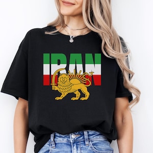 Op de afbeelding: Zwart T-shirt met het woord "IRAN" in groene en witte blokletters. Daaronder een gouden leeuw met een zwaard, met een zon erachter. Het shirt wordt gedragen met een blauwe spijkerbroek.