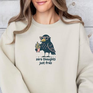 May include: Cream-colored sweatshirt with a graphic of a cartoon crow wearing sunglasses and holding fries. The text "zero thoughts just fries" is below the crow. The sweatshirt is a casual style.