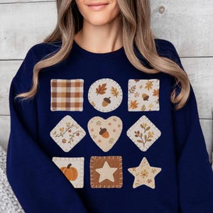 Peut inclure: Un sweat-shirt bleu marine orné de neuf écussons sur le thème de l'automne. Les écussons comprennent un carré à carreaux, des feuilles, des glands, des citrouilles et des étoiles. Le sweat-shirt est porté par une personne assise sur un canapé.