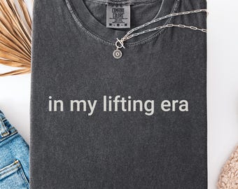 Può includere: Una maglietta grigio scuro con la scritta bianca "in my lifting era". La maglietta piegata è appoggiata su una superficie bianca, accanto a jeans blu, scarpe da ginnastica bianche e una collana d'argento. È presente anche una foglia di palma secca.