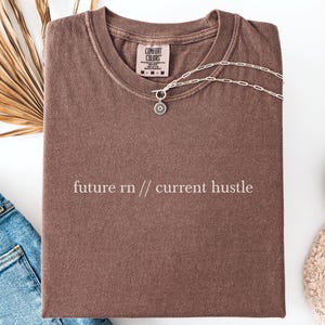 May include: A brown t-shirt with the text "future rn // current hustle" is laid flat, with a silver necklace draped over it. The shirt is next to a pair of blue jeans, white trainers, and a dried palm frond.