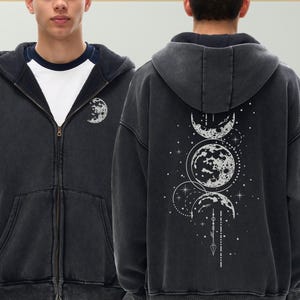 Puede incluir: Sudadera con capucha y cremallera gris oscuro con un diseño de fases lunares blancas en la espalda y un gráfico de luna más pequeño en la parte delantera. La sudadera tiene capucha y un ajuste relajado. El diseño incluye estrellas y elementos celestiales.