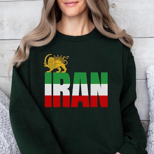 Könnte beinhalten: Dunkelgrünes Sweatshirt mit dem Wort "IRAN" in weißen, grünen und roten Streifen. Über dem Wort befindet sich ein goldenes Löwenemblem. Das Sweatshirt ist ein lässiger Stil.