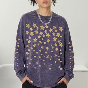 Può includere: Una felpa viola a maniche lunghe con un motivo floreale. Il design presenta numerosi fiori gialli sparsi sul davanti e sulle maniche. La felpa ha un aspetto vintage e una vestibilità rilassata.