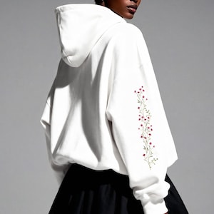 May include: A white hoodie with a floral embroidery design on the left sleeve. The hoodie is paired with a black skirt. The model is wearing multiple rings. The background is a solid gray.