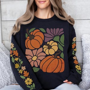 May include: Black sweatshirt featuring a fall-themed design with orange pumpkins, flowers, and leaves. The sleeves have floral patterns in orange, yellow, and green. The sweatshirt is a cozy, seasonal apparel item.