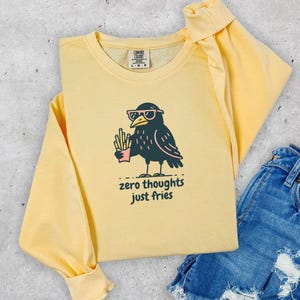 May include: A light yellow long-sleeve sweatshirt with a graphic of a black bird wearing sunglasses and holding fries. The text "zero thoughts just fries" is below the bird. The sweatshirt is paired with denim shorts.