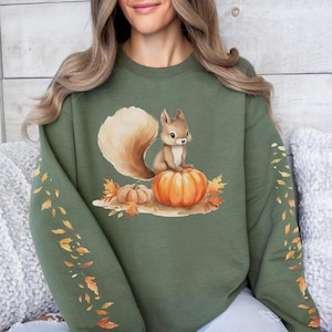 May include: Olive green sweatshirt featuring a squirrel sitting on a pumpkin with fall leaves. The sweatshirt has a crew neck and long sleeves with a leaf design. The design is in shades of brown, orange, and tan.