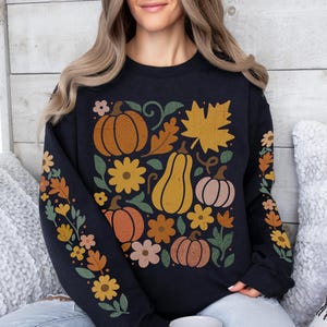 May include: Black sweatshirt with a fall-themed design featuring pumpkins, flowers, and leaves in shades of orange, yellow, and brown. The sleeves have floral patterns. The design is centered on the chest.