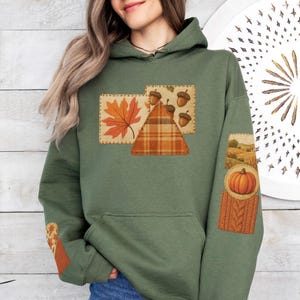 May include: An olive green hoodie with autumn-themed patches. Patches include a maple leaf, acorns, a plaid triangle, and a pumpkin with a cable knit sleeve. The hoodie is made of a soft material and has a hood and a front pocket.