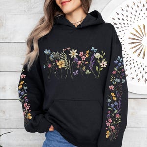 Pode incluir: Moletom preto com capuz com um design floral no peito e nas mangas. O padrão floral apresenta flores coloridas em tons de azul, amarelo, rosa e roxo. O moletom tem um bolso frontal e um capuz com cordão.
