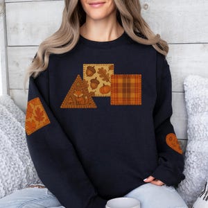 May include: A navy blue sweatshirt with autumn-themed patches. The patches feature a fox, acorns, pumpkins, and plaid patterns in shades of orange, brown, and yellow. The sweatshirt has matching elbow patches with leaf designs.