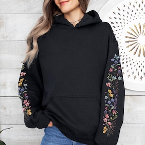 May include: Black hoodie with floral embroidery on the sleeves. The embroidery features a variety of colourful flowers, including pink, yellow, blue, and purple. The hoodie has a front pocket and a drawstring hood.