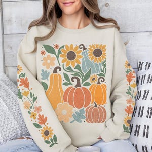May include: Cream-colored sweatshirt featuring a fall-themed design. The design includes sunflowers, pumpkins, and floral elements in shades of orange, yellow, and blue. The sleeves have a floral pattern. The sweatshirt is a cozy, seasonal apparel item.