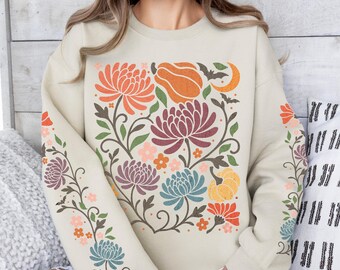 May include: Beige sweatshirt with a floral design featuring orange, purple, and blue flowers, a pumpkin, and a crescent moon. The sleeves have matching floral patterns. A white coffee cup is in the foreground.