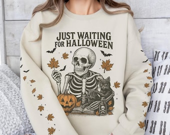 May include: Cream-colored sweatshirt with the text "JUST WAITING FOR HALLOWEEN" above a skeleton illustration. The skeleton is holding a leaf and sitting with a black cat and a jack-o'-lantern. The sleeves have a pattern of leaves and bats.