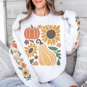 May include: White sweatshirt featuring a fall-themed design. The design includes an orange pumpkin, a sunflower, and a gourd, along with floral accents. The sleeves are decorated with additional floral patterns in autumn colors. The sweatshirt is a comfortable, casual style.
