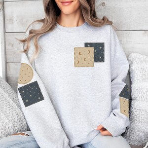Puede incluir: Una sudadera gris claro con parches de diseños celestiales. Los parches incluyen lunas crecientes, estrellas y constelaciones en color beige y azul marino. La sudadera tiene cuello redondo y puños y dobladillo acanalados.