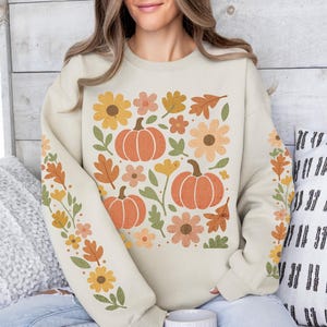 May include: Cream-colored sweatshirt featuring a fall-themed floral design with pumpkins, flowers, and leaves in shades of orange, yellow, and brown. The sleeves have a similar floral pattern. The sweatshirt is a cozy, seasonal apparel item.