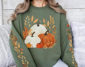 Könnte beinhalten: Olivgrünes Sweatshirt mit herbstlichem Design. Die Grafik zeigt drei Kürbisse in Weiß und Orange, umgeben von Herbstblättern. Die Ärmel sind mit einem Blättermuster verziert.