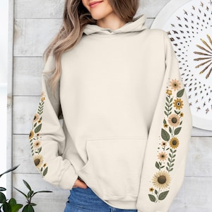Peut inclure: Sweat à capuche crème avec une poche avant et un motif floral sur les manches. Les manches présentent un motif vertical de tournesols, de feuilles et de petites fleurs dans les tons de jaune, marron et vert. Le sweat à capuche est en matière douce.