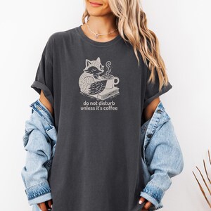 May include: Dark gray t-shirt featuring a whimsical fox illustration with a coffee cup and book. The text "do not disturb unless it's coffee" is printed below the graphic. The shirt has rolled-up sleeves and a relaxed fit.