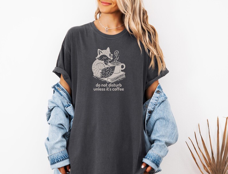 May include: Dark gray t-shirt featuring a whimsical fox illustration with a coffee cup and book. The text "do not disturb unless it's coffee" is printed below the graphic. The shirt has rolled-up sleeves and a relaxed fit.