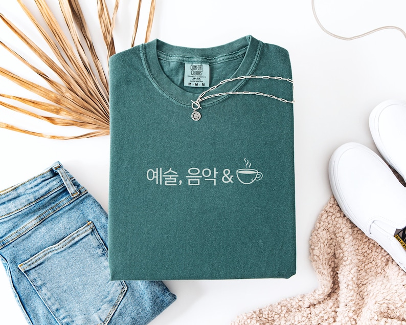 Peut inclure: Un t-shirt Comfort Colors vert d'eau avec du texte cor&eacute;en blanc et un dessin de tasse &agrave; caf&eacute;. Un collier en argent est pos&eacute; sur le t-shirt, &agrave; c&ocirc;t&eacute; d'un jean bleu et de chaussures blanches &agrave; enfiler. Une feuille de palmier s&eacute;ch&eacute;e est &eacute;galement dans l'image.