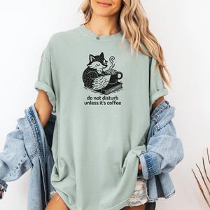 May include: Sage green t-shirt with a black and white graphic of a fox curled up with a coffee cup and a book. The text "do not disturb unless it's coffee" is below the graphic. The shirt is a relaxed fit.