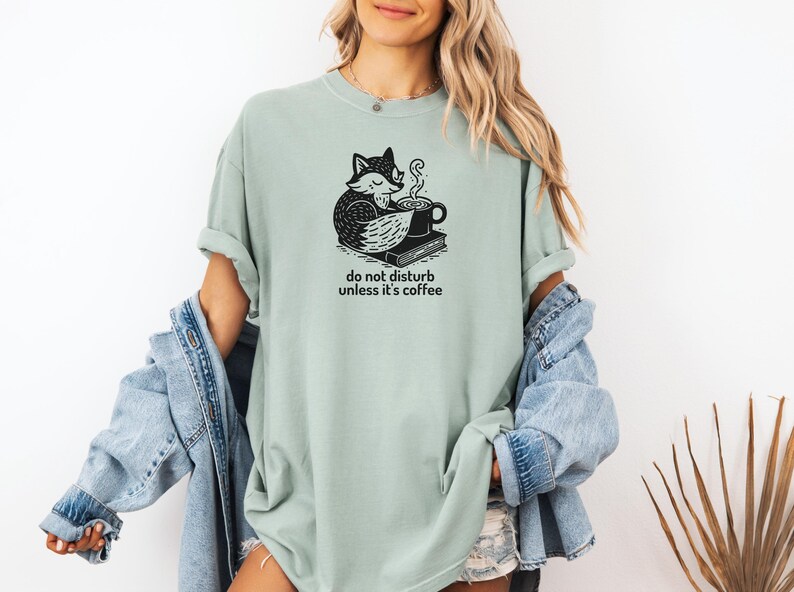 May include: Sage green t-shirt with a black and white graphic of a fox curled up with a coffee cup and a book. The text "do not disturb unless it's coffee" is below the graphic. The shirt is a relaxed fit.