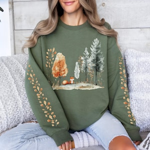 Pode incluir: Um moletom verde-oliva com uma cena de floresta em aquarela e uma raposa. O design inclui &aacute;rvores em tons de laranja, branco e verde. As mangas t&ecirc;m um padr&atilde;o de folhas repetido em laranja e dourado.