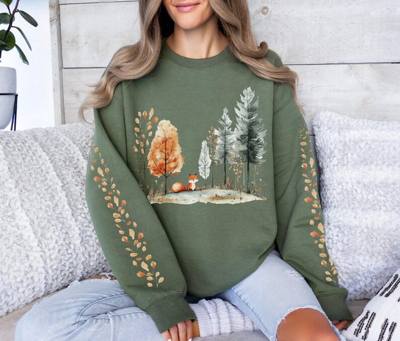 Pode incluir: Um moletom verde-oliva com uma cena de floresta em aquarela e uma raposa. O design inclui &aacute;rvores em tons de laranja, branco e verde. As mangas t&ecirc;m um padr&atilde;o de folhas repetido em laranja e dourado.