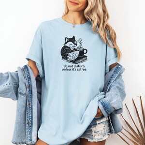 May include: Light blue t-shirt with a black and white graphic of a fox curled up with a coffee cup and book. The text "do not disturb unless it's coffee" is below the graphic. The shirt is made of a soft material.
