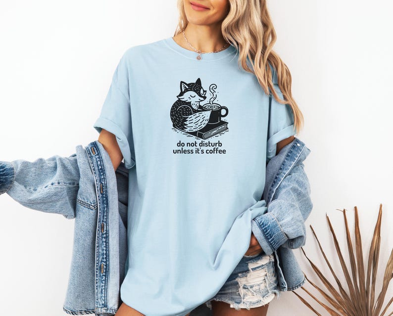 May include: Light blue t-shirt with a black and white graphic of a fox curled up with a coffee cup and book. The text "do not disturb unless it's coffee" is below the graphic. The shirt is made of a soft material.