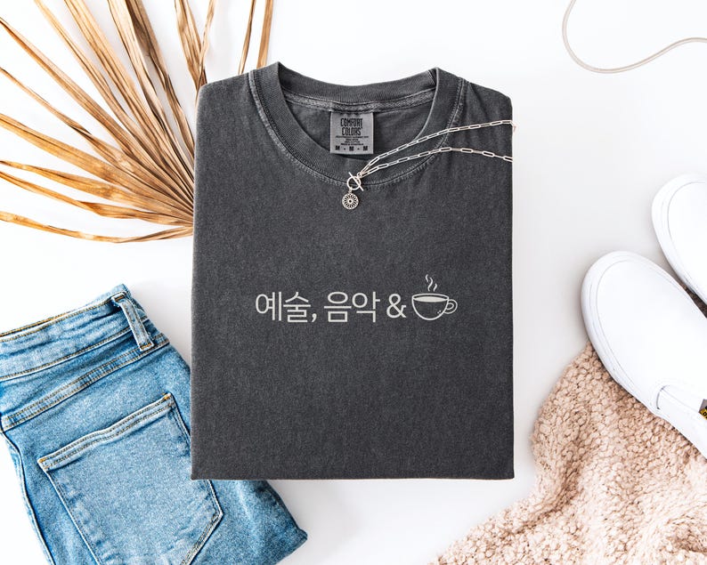 Peut inclure: Un t-shirt gris anthracite Comfort Colors avec du texte cor&eacute;en blanc et un dessin de tasse &agrave; caf&eacute;. Le t-shirt est associ&eacute; &agrave; un jean bleu, des chaussures blanches &agrave; enfiler et un collier argent&eacute;. Une feuille de palmier s&eacute;ch&eacute;e ajoute un &eacute;l&eacute;ment naturel.
